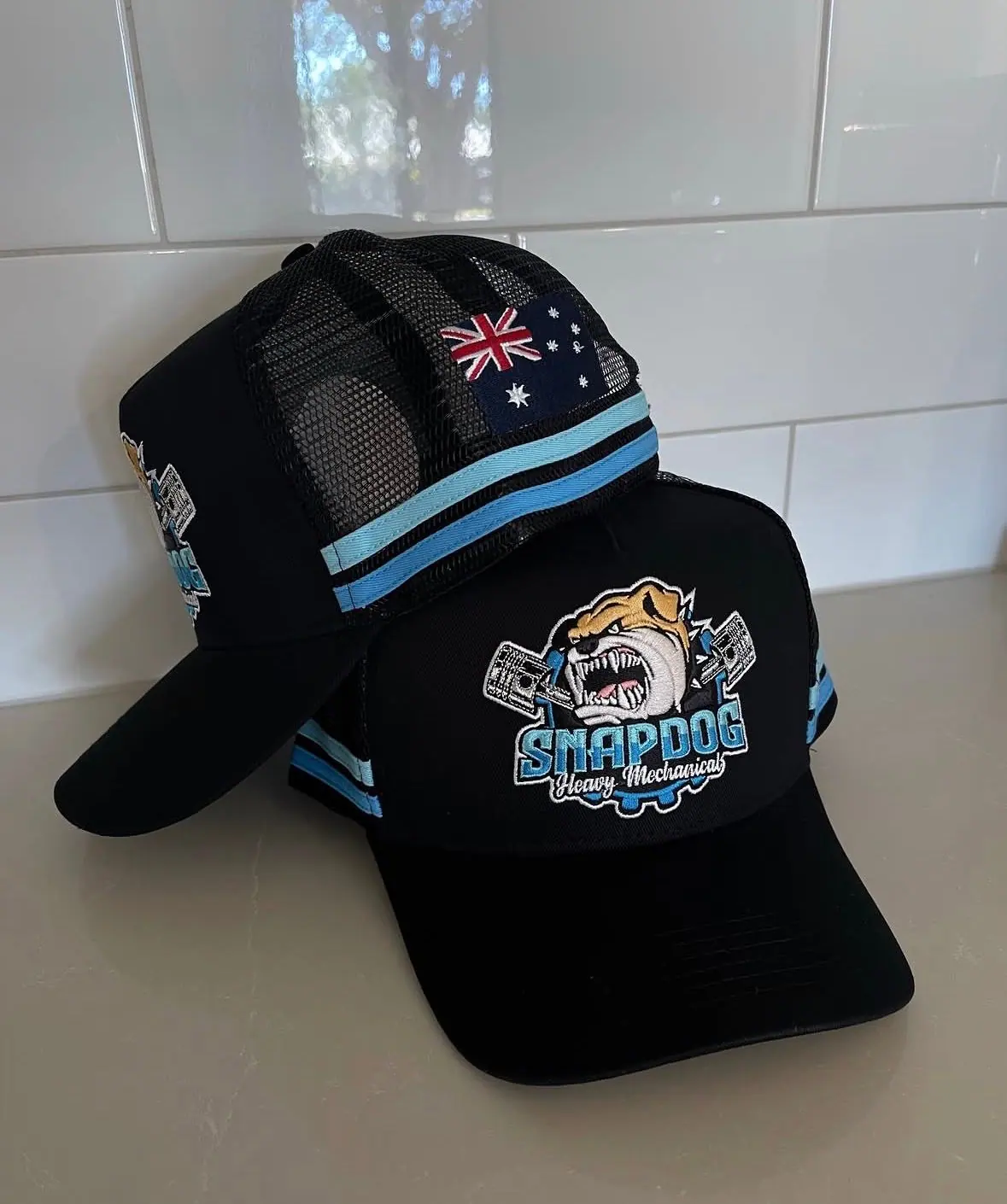 Two black SnapDog trucker caps with the embroidered bulldog logo. One shows the light blue striped band and an Australian flag on the mesh side.