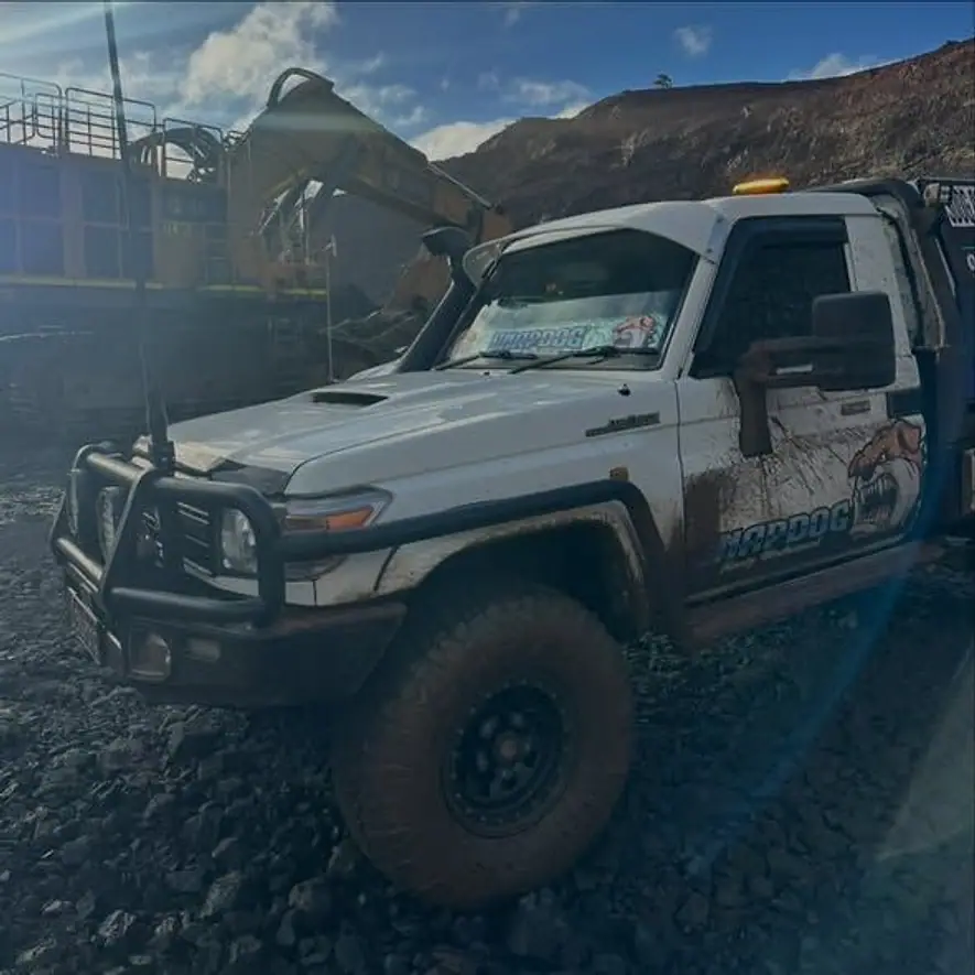 SnapDog's tray-back parked on rough ground below a mine pit wall with an excavator behind and low sun flaring across the lens.